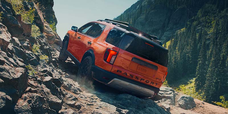 Orange SUV climbing a rocky mountain trail with forested cliffs in the background.